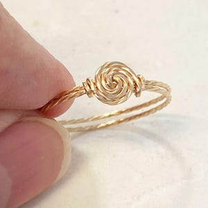 May include: A gold-colored wire ring featuring a central spiral knot design. The band is constructed from two twisted strands of wire that meet at the center to form a circular, coiled focal point, creating a continuous, textured metallic appearance.