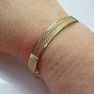 K&ouml;nnte beinhalten: Ein goldfarbenes Drahtarmband mit geflochtenem Muster. Das Armband befindet sich an einem Handgelenk.