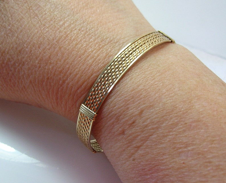 K&ouml;nnte beinhalten: Ein goldfarbenes Drahtarmband mit geflochtenem Muster. Das Armband befindet sich an einem Handgelenk.