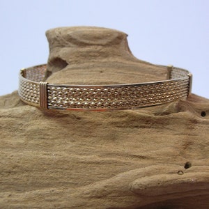 K&ouml;nnte beinhalten: Ein goldfarbenes Metallarmband mit geflochtenem Design. Das Armband wird auf einem St&uuml;ck Holz pr&auml;sentiert.