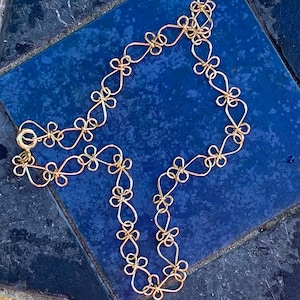 May include: A gold-coloured necklace featuring a repeating floral pattern. The necklace is crafted from thin, looped wire, giving it a light and airy feel. It has a simple gold-coloured hook and eye clasp. The necklace is displayed on a dark blue tiled surface.