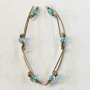 May include: A pair of gold-toned earrings with a unique design. Each earring features a curved wire frame adorned with light blue, oval-shaped beads and small gold-colored beads. The earrings have a delicate and elegant appearance.