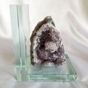 May include: A clear glass bookend featuring a jagged amethyst geode with deep purple crystalline structures embedded within a grey and white rocky exterior, mounted on a flat, transparent glass base with a vertical support panel.