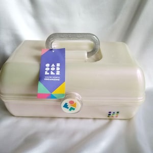May include: A cream-colored plastic storage case with a glittery translucent handle. A purple tag hangs from the handle with the text CABOODLES YOU'RE WORTH ORGANIZING. A circular latch features a geometric logo, and the brand name is printed on the front.