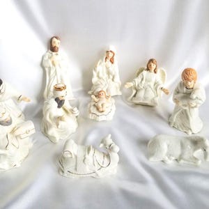 May include: A set of white porcelain nativity figurines featuring Mary, Joseph, the baby Jesus in a manger, an angel, three wise men, a camel, and a donkey. Each figure is accented with delicate gold trim and painted facial features.