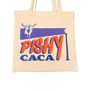 May include: A beige canvas tote bag with a purple and blue design. The design features two figures dancing on a purple banner with the text "PISHY CACA" in orange and blue.