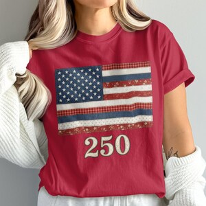 Może przedstawiać: Czerwony t-shirt z krótkim rękawem z grafiką flagi amerykańskiej złożoną z różnych wzorzystych pasków materiału, w tym w kwiaty i kratę. Pod flagą znajduje się nadruk liczby 250 w kremowym kolorze z ciemnym obrysem.