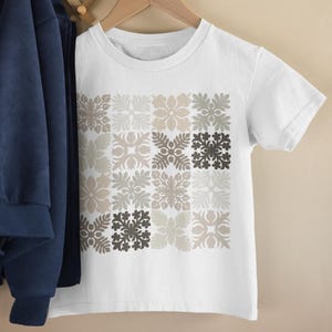 May include: White t-shirt with a repeating floral pattern in shades of gray and beige. The short-sleeved shirt is hanging on a wooden hanger. A navy blue jacket and a stuffed elephant are visible on a wooden wall rack.