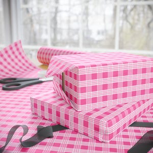 Könnte beinhalten: Zwei rosa und weiße Gingham-Geschenkboxen, die übereinander gestapelt sind. Die Boxen sind in rosa und weißes Gingham-Papier eingewickelt. Ein schwarzes Band ist um die untere Box gebunden.