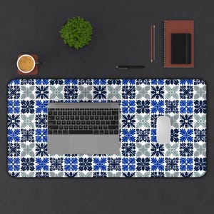 May include: A desk mat featuring a blue, white, and grey floral tile design. A laptop, mouse, and keyboard are on the mat. A small plant, coffee cup, pen, pencil, and notebook are also on the desk.