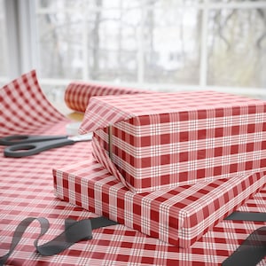 May include: Two red and white gingham gift boxes stacked on top of each other. The boxes are wrapped in red and white checkered paper. A pair of scissors and a roll of wrapping paper are in the background.
