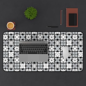 May include: A black desk mat with a white and gray floral pattern, a laptop, and a white mouse. A cup of coffee, a small plant, a pen, a pencil, and a notebook are also on the desk.