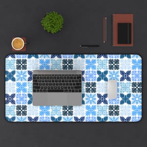 May include: A rectangular desk mat featuring a blue and white floral tile design. A laptop, mouse, coffee cup, pen, notebook, and small plant are on the mat. The mat is on a dark surface.