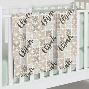 May include: A baby blanket with a repeating floral pattern in shades of beige, tan, and white. The name "Elijah" is written in a cursive font across the blanket. The blanket is draped over a white crib.