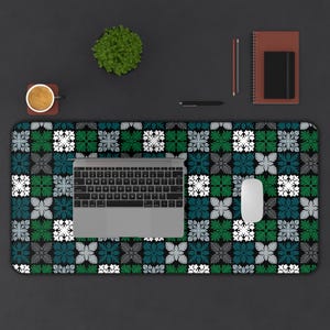 May include: A desk mat with a floral pattern in green, teal, grey, and white on a black background. A laptop, mouse, notebook, pen, and a small plant are on the mat. The mat is approximately 70 cm long.