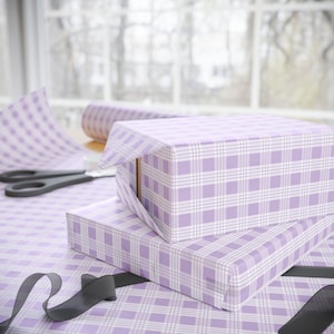 May include: Two gift boxes wrapped in lavender and white plaid wrapping paper. A roll of matching paper, scissors, and a black ribbon are also visible, suggesting a gift-wrapping scene.