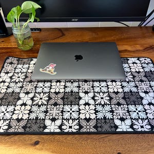 May include: A black laptop with an Apple logo sits on a patterned mousepad. The mousepad features a repeating floral design in black, white, and gray. A sticker is on the laptop. A monitor and a plant in a jar are in the background.