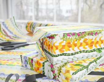Könnte beinhalten: Zwei Geschenkboxen, die in weißes Papier mit einem Blumenmuster aus gelben, orangefarbenen, rosa und grünen Blumen eingewickelt sind. Die Boxen sind übereinander gestapelt und von einem schwarzen Band umgeben.