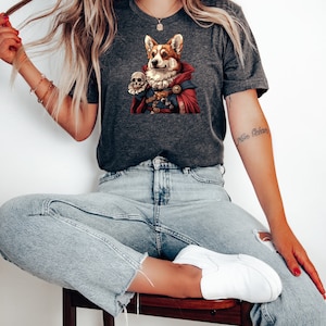 May include: A woman wearing a gray t-shirt with a graphic of a corgi dressed as a wizard holding a skull. The corgi is wearing a red cape and has a gold chain around its neck. The woman is also wearing blue jeans and white sneakers.