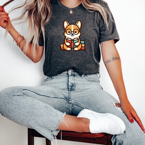 May include: A woman wearing a dark grey t-shirt with a cartoon image of a corgi wearing glasses and reading a book. The corgi is sitting on a stack of books. The woman is wearing light blue jeans and white trainers.