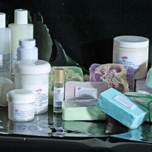 May include: Assortment of bath and body products including soaps, lotions, and essential oils. The products are arranged on a black surface. The soaps are in various colors and shapes, including a lavender soap with a loofah sponge.