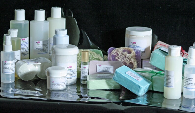 May include: Assortment of bath and body products including soaps, lotions, and essential oils. The products are arranged on a black surface. The soaps are in various colors and shapes, including a lavender soap with a loofah sponge.