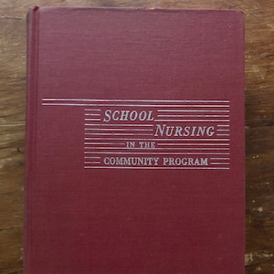 May include: A maroon hardback book with the title "School Nursing in the Community Program" printed in silver lettering.