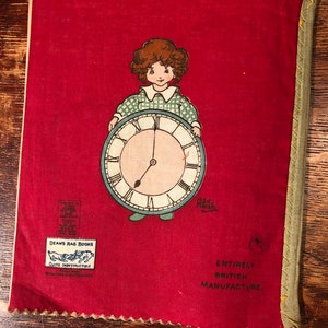 Puede incluir: Una cubierta de libro de tela roja con una ni&ntilde;a sosteniendo un reloj. La cubierta del libro tiene el texto "Dean's Rag Books, Quite Indestructible, Registered in all Countries" y "Entirely British Manufacture".