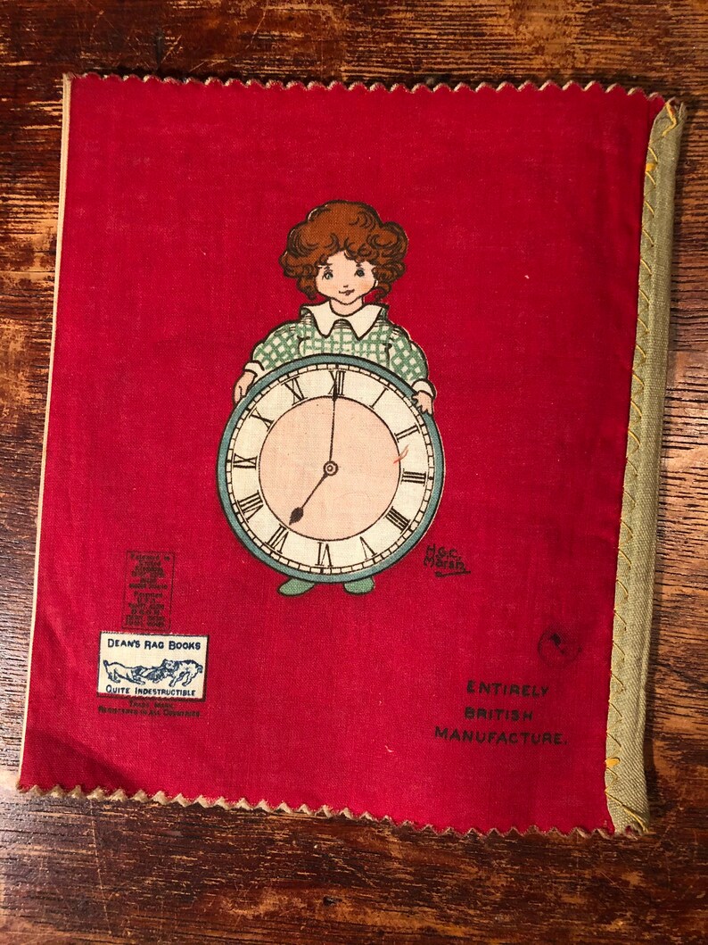 Puede incluir: Una cubierta de libro de tela roja con una ni&ntilde;a sosteniendo un reloj. La cubierta del libro tiene el texto "Dean's Rag Books, Quite Indestructible, Registered in all Countries" y "Entirely British Manufacture".