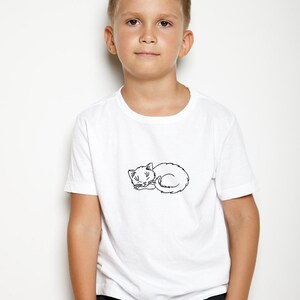 Puede incluir: Una persona joven con una camiseta blanca con un contorno negro de un gato durmiendo en la parte delantera. La persona lleva pantalones negros y tiene el pelo corto casta&ntilde;o.