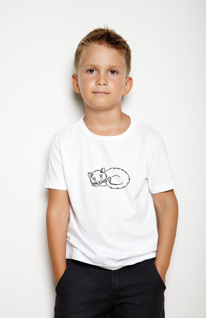 Puede incluir: Una persona joven con una camiseta blanca con un contorno negro de un gato durmiendo en la parte delantera. La persona lleva pantalones negros y tiene el pelo corto casta&ntilde;o.