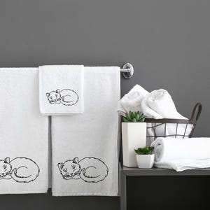Puede incluir: Tres toallas de ba&ntilde;o blancas con dise&ntilde;os de gatos durmiendo bordados. Las toallas est&aacute;n colgadas en un toallero plateado. Hay toallas blancas adicionales en una cesta de alambre y en un estante.