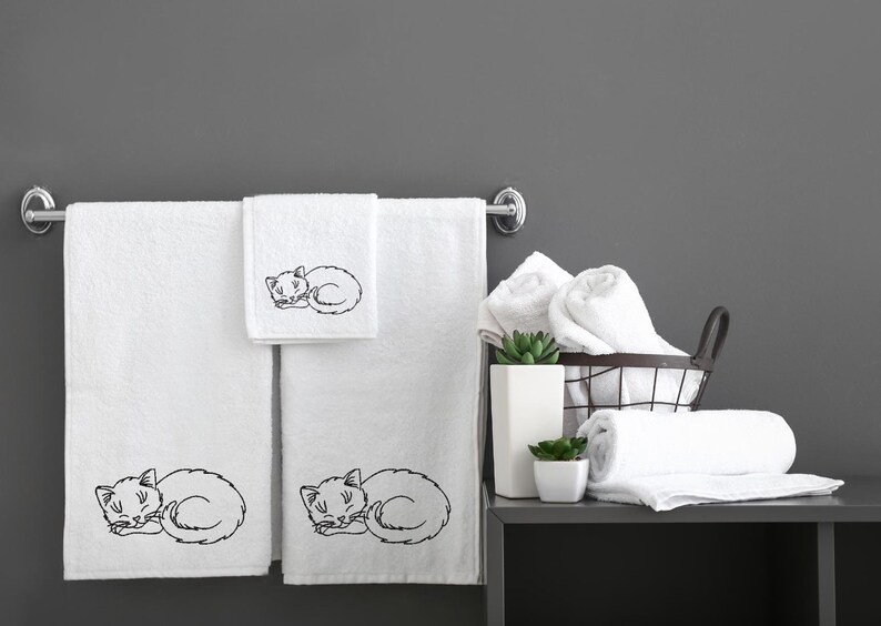 Puede incluir: Tres toallas de ba&ntilde;o blancas con dise&ntilde;os de gatos durmiendo bordados. Las toallas est&aacute;n colgadas en un toallero plateado. Hay toallas blancas adicionales en una cesta de alambre y en un estante.