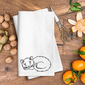 Puede incluir: Una toalla de cocina blanca con un dise&ntilde;o bordado en negro de un gato durmiendo. La toalla est&aacute; doblada y puesta sobre una superficie de madera con nueces y naranjas.