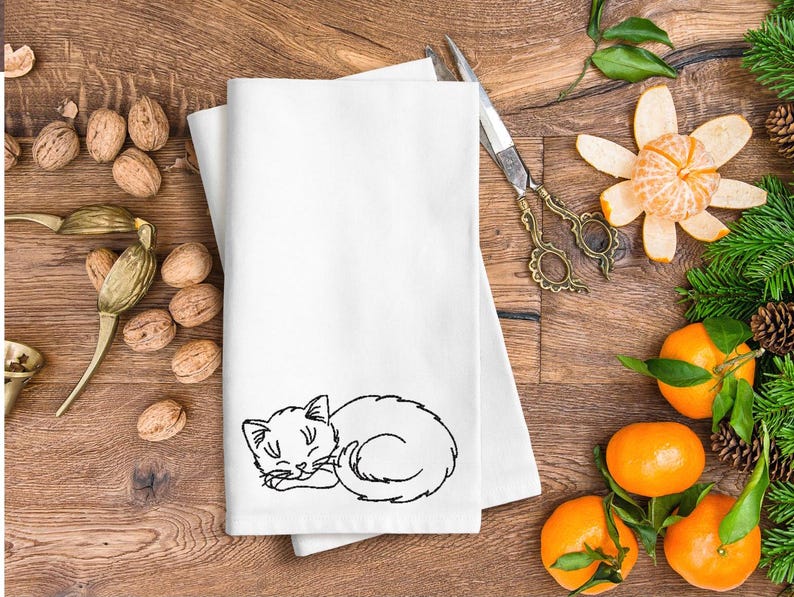 Puede incluir: Una toalla de cocina blanca con un dise&ntilde;o bordado en negro de un gato durmiendo. La toalla est&aacute; doblada y puesta sobre una superficie de madera con nueces y naranjas.
