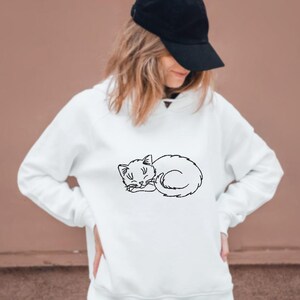 Puede incluir: Una sudadera blanca con un contorno negro de un gato durmiendo en la parte delantera.