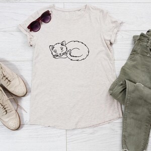 Puede incluir: Una camiseta beige con un dibujo de l&iacute;nea negra de un gato durmiendo. La camiseta est&aacute; colocada sobre un fondo de madera blanca con un par de zapatos beige y un par de pantalones vaqueros verdes.