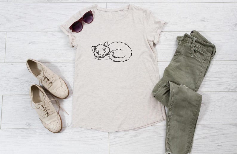 Puede incluir: Una camiseta beige con un dibujo de l&iacute;nea negra de un gato durmiendo. La camiseta est&aacute; colocada sobre un fondo de madera blanca con un par de zapatos beige y un par de pantalones vaqueros verdes.