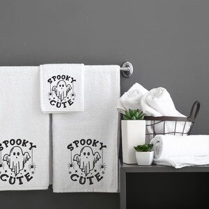 May include: Three white bath towels with black embroidered text that reads "Spooky Cute" and a ghost graphic. The towels are hanging on a chrome towel rack. There are additional white towels in a wire basket on a grey shelf.