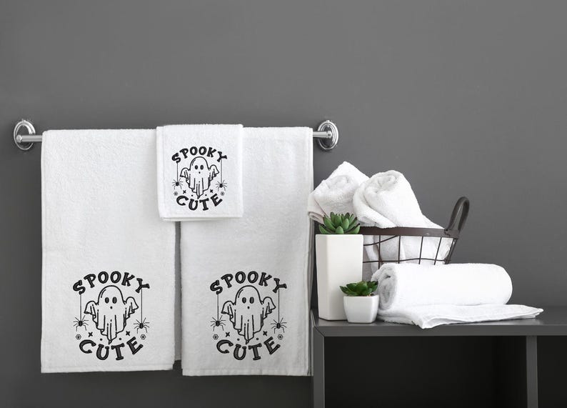 May include: Three white bath towels with black embroidered text that reads "Spooky Cute" and a ghost graphic. The towels are hanging on a chrome towel rack. There are additional white towels in a wire basket on a grey shelf.