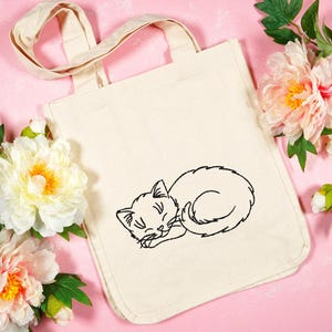 Puede incluir: Una bolsa de lona blanca con una imagen bordada en negro de un gato durmiendo. La bolsa tiene dos asas y est&aacute; sobre un fondo rosa con flores rosas.