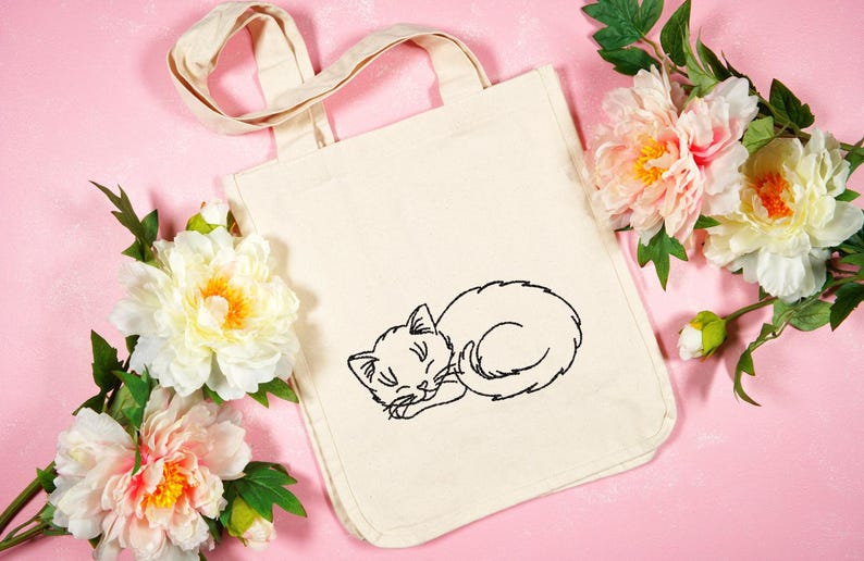 Puede incluir: Una bolsa de lona blanca con una imagen bordada en negro de un gato durmiendo. La bolsa tiene dos asas y est&aacute; sobre un fondo rosa con flores rosas.