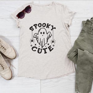 May include: A beige t-shirt with a black graphic of a ghost with the text "Spooky Cute" on it. The shirt is laid out on a white wooden surface with a pair of brown sunglasses, a pair of beige shoes, and a pair of green jeans.