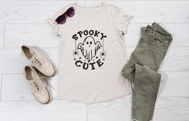 May include: A beige t-shirt with a black graphic of a ghost with the text "Spooky Cute" on it. The shirt is laid out on a white wooden surface with a pair of brown sunglasses, a pair of beige shoes, and a pair of green jeans.