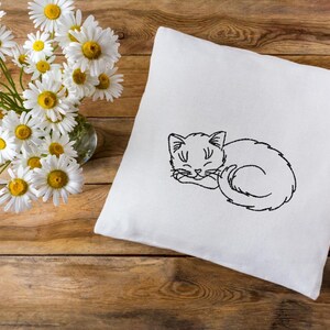 Puede incluir: Una almohada blanca con un dibujo de l&iacute;nea negra de un gato durmiendo bordado en ella. La almohada est&aacute; sobre una superficie de madera con un ramo de margaritas blancas en un jarr&oacute;n de vidrio.