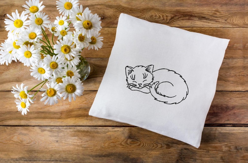 Puede incluir: Una almohada blanca con un dibujo de l&iacute;nea negra de un gato durmiendo bordado en ella. La almohada est&aacute; sobre una superficie de madera con un ramo de margaritas blancas en un jarr&oacute;n de vidrio.
