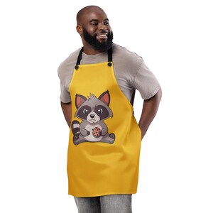 May include: A yellow apron featuring a cartoon raccoon holding a cookie. The raccoon is gray and white with black accents. The apron has black straps and buttons.