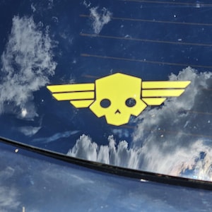 May include: A bright yellow vinyl decal featuring a stylized skull with a geometric, hexagonal cranium and two sets of horizontal, tiered wings extending from the sides, applied to a dark blue surface reflecting clouds and sky.