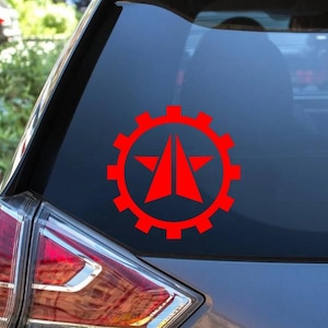 May include: A bright red vinyl decal applied to a vehicle window. The design features a central five-pointed star split vertically by a thin line, enclosed within a circular gear shape with prominent teeth along the outer edge of the graphic.