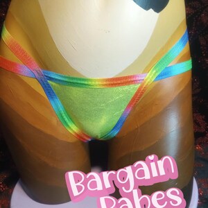 Puede incluir: Un torso de maniquí pintado con tonos de piel lleva un bikini verde y con ribetes de arcoíris. El bikini tiene un brillo metálico. El texto "Bargain Babes" se muestra en una pegatina rosa en la base del maniquí.